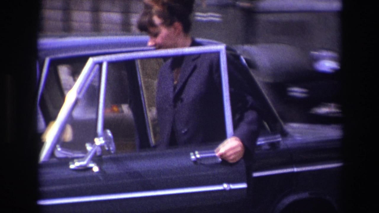 Woman holding the open rear door of a black sedan beside a stone building.