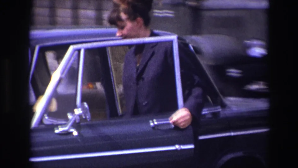 A woman in a dark outfit holds the open door of a classic black sedan in a 1967 France home-movie frame.