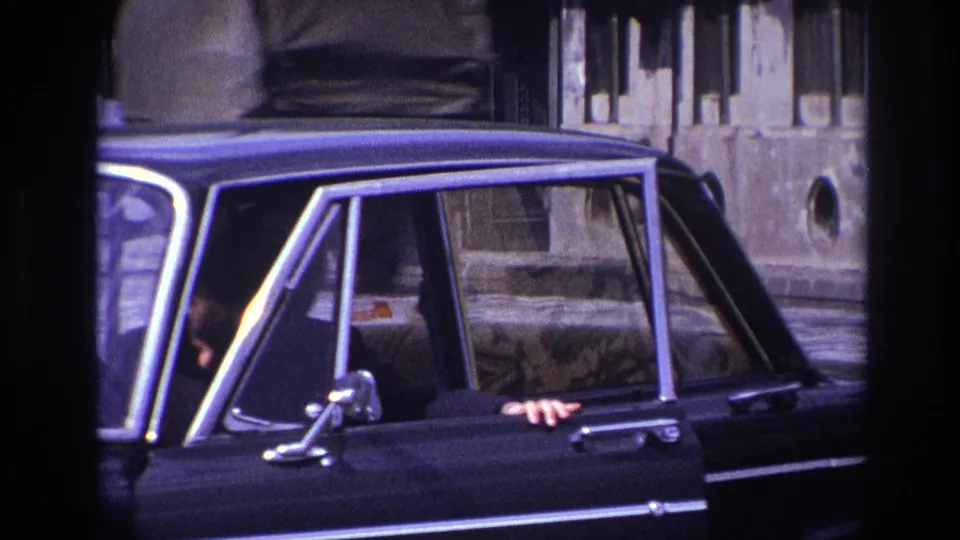 The woman is mostly inside the black sedan, with her hand and the open door still visible against the stone street backdrop.