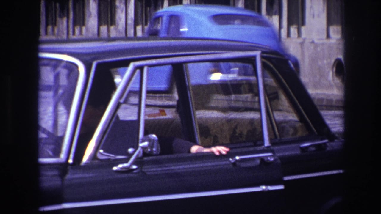 1967 France 8mm frame showing a black sedan parked or moving along a street, with a hand resting near the side window.