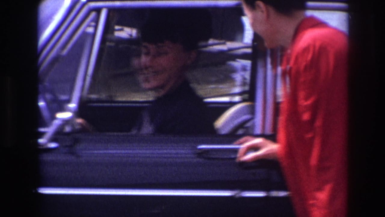 1967 France street view of a smiling woman in a dark hat inside a black sedan while a man in a red jacket leans near the window.