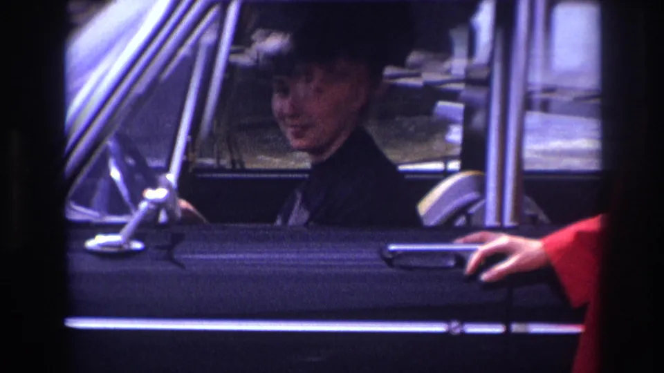 1967 France 8mm frame with a woman seated inside a black sedan, smiling as a red-sleeved hand reaches toward the door.