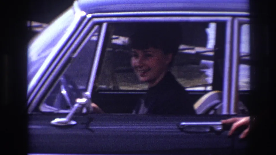 1967 France frame of a smiling woman in dark clothing inside a black sedan, seen through the side window with a hand near the door.