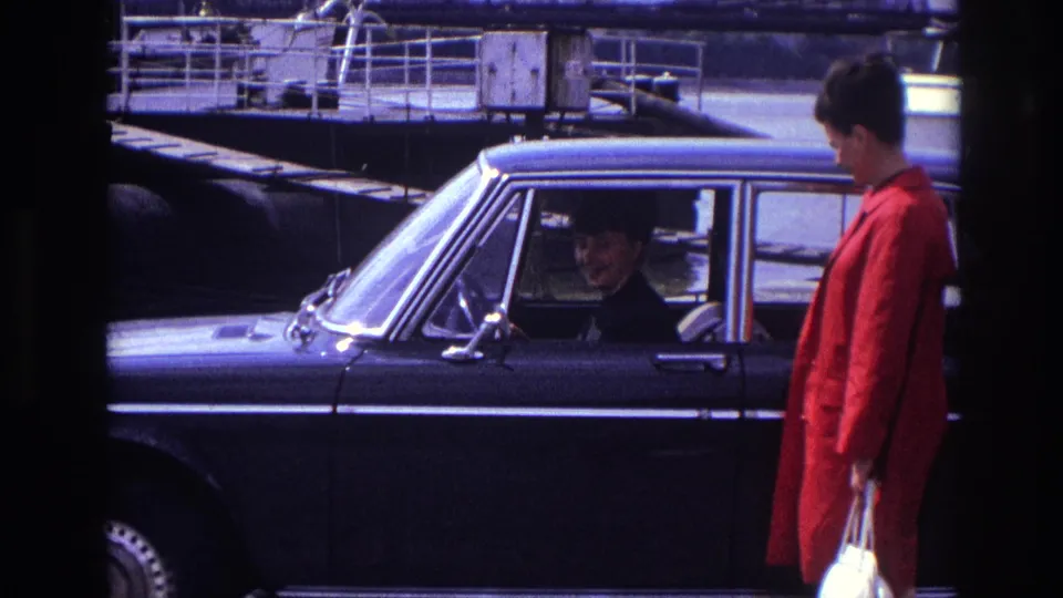 A man in a red coat holding a white bag stands beside the black car while the woman remains inside.