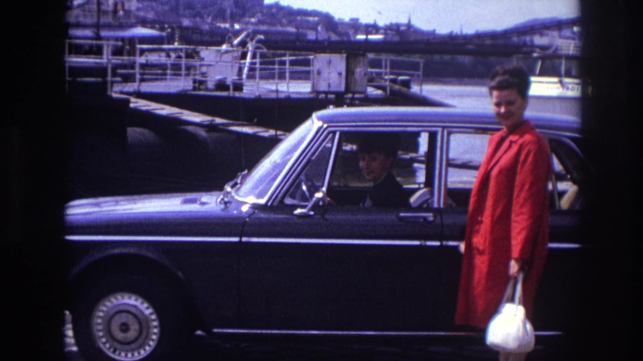 1967 France home-movie frame of a woman in a red coat with a white handbag beside a dark sedan near a riverside quay.
