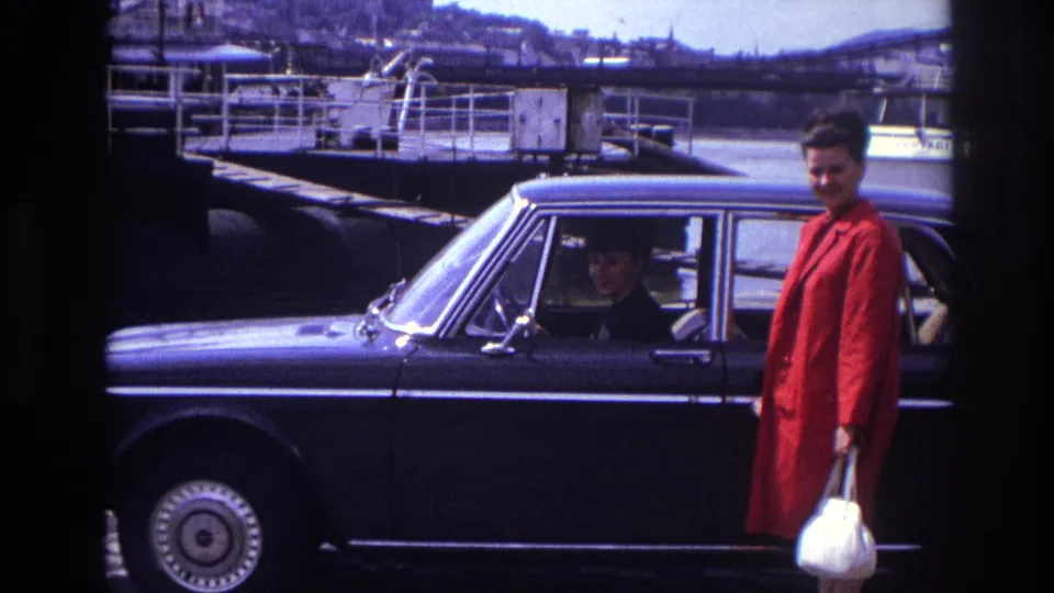 1967 France home-movie frame of a woman in a red coat with a white handbag beside a dark sedan near a riverside quay.
