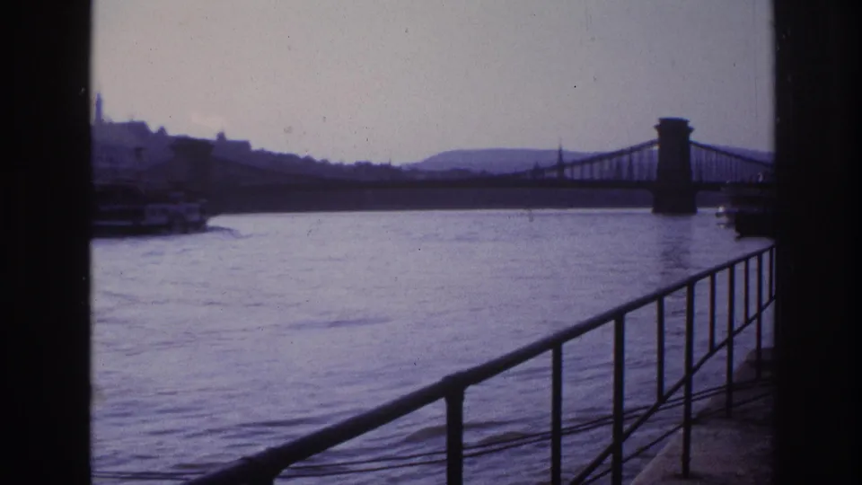 1967 France 8mm frame with river railing in the foreground, a suspension bridge, and a ferry partly leaving view.