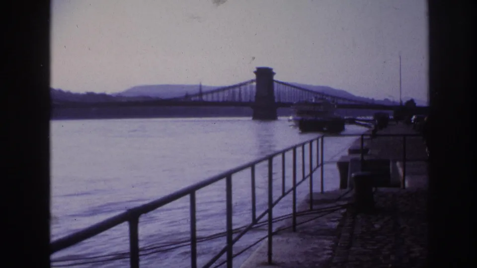1967 8mm travel frame showing a waterfront railing, broad river or harbor, distant suspension bridge, and shadowed bridge approach in France metadata.
