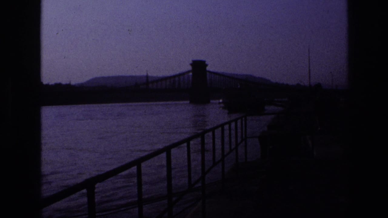 Darker waterfront view with railing, water, and a suspension bridge silhouetted across the frame.