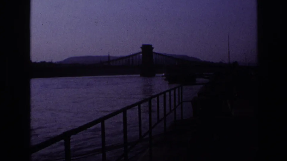 Underexposed 1967 home-movie frame of a riverside walkway and suspension bridge, with the water and distant hills visible through purple color shift.