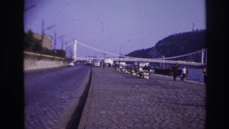 1967 travel footage frame showing a cobblestone promenade or road beside the water, with pedestrians, cars, bridge structure, and hillside in the background.