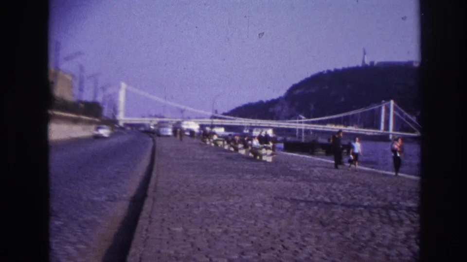 1967 8mm frame of a public waterfront road in France metadata, with distant pedestrians, parked cars, suspension bridge spans, and a tree-covered hill.