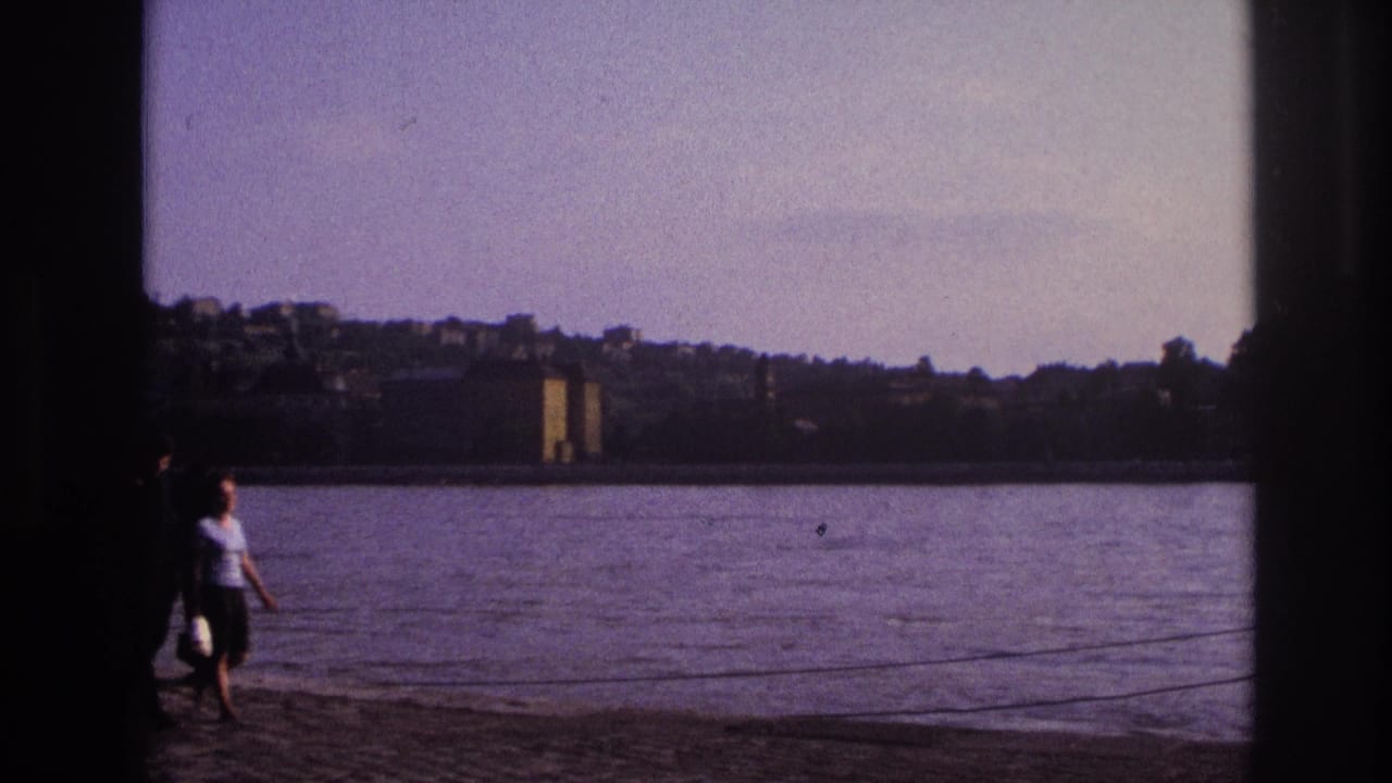Two adults walk along a paved lakeside path with water and distant buildings behind them.
