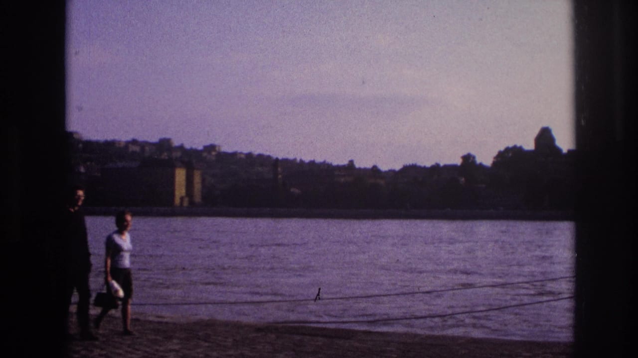 A woman in a light top walks along a French lakeside path as another adult remains partly obscured by dark framing.