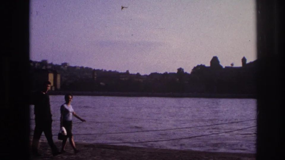 Two adults move along the lakeside promenade in 1967 France, backed by water, distant buildings, and shadowed framing.