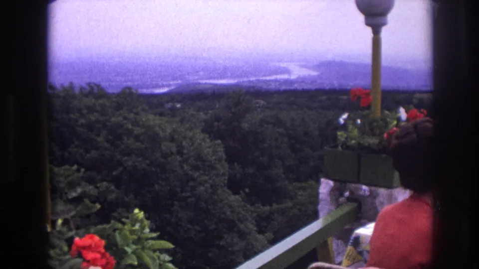 1967 France 8mm frame with a woman in red seated on a balcony overlooking trees and distant water.