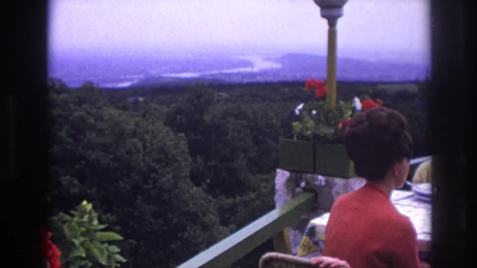 1967 France home-movie frame showing a woman in red seated at a balcony table above trees and water.