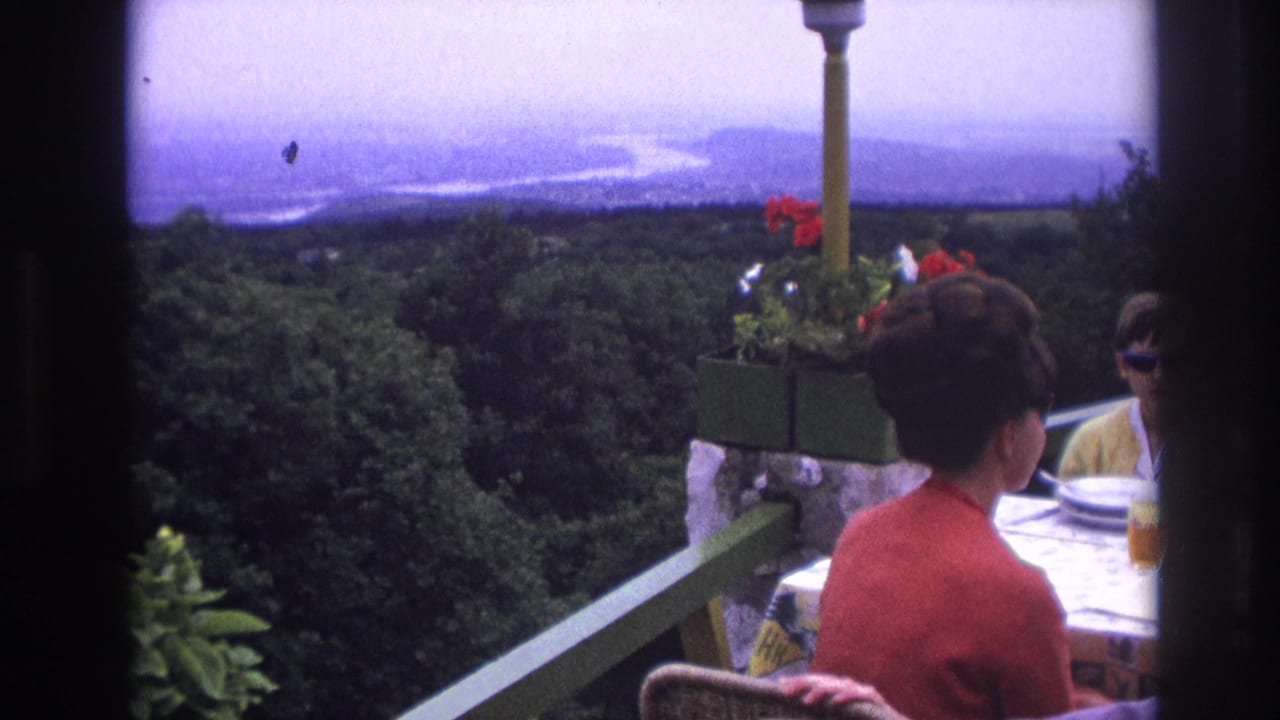 1967 France home-movie frame showing a woman and child at a terrace restaurant table overlooking forest and a winding river.