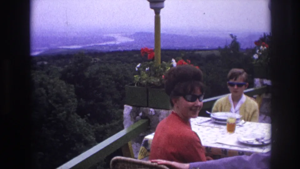 1967 France home-movie frame of a woman smiling in sunglasses at an outdoor table, with a child seated behind and landscape beyond.