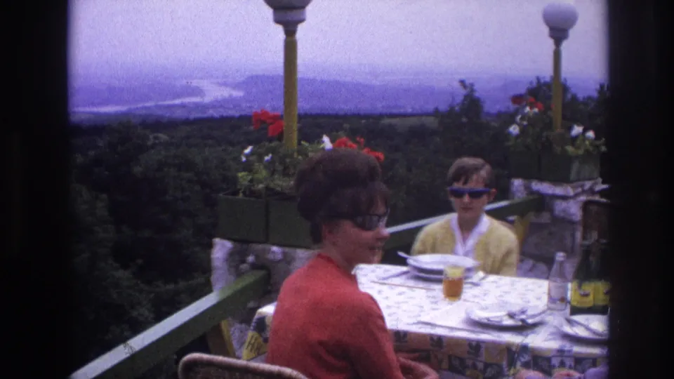 1967 France terrace restaurant frame with woman and child at a patterned table overlooking dense trees and a hazy river landscape.