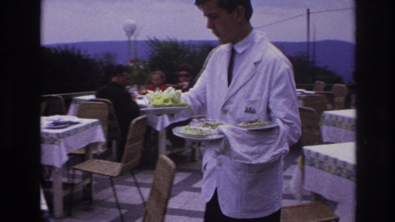 In France in 1967, a waiter in a white jacket and tie moves between terrace tables while carrying plated salads.