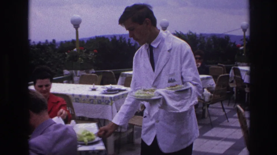 The waiter serves food to diners at a French outdoor restaurant terrace, with hedges, lamps, and hills behind the tables.