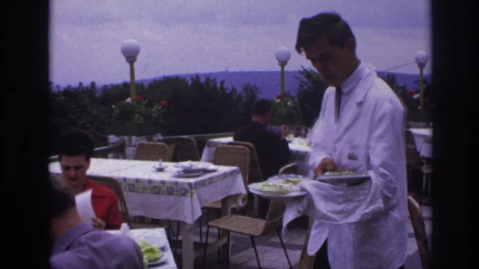 A waiter in a white service jacket holds salad plates beside seated diners on a 1967 French restaurant terrace.
