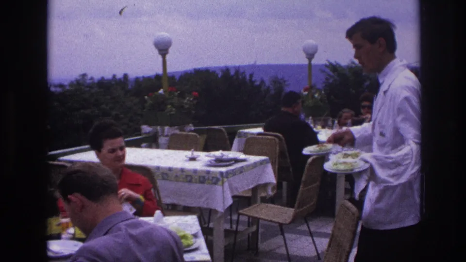 At the end of the 1967 France clip, the waiter stands near diners on a terrace as plated food and distant hills remain visible.