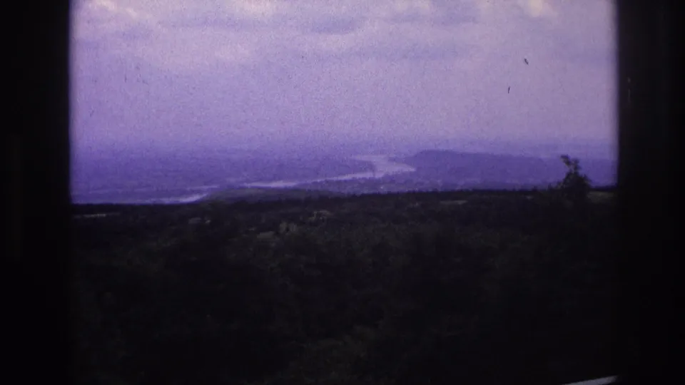 Wide 1967 France travel-footage frame showing an overcast river landscape and hills through dark architectural framing.