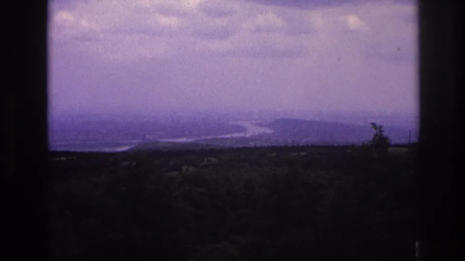 1967 France home-movie frame showing a hazy river valley and wooded overlook with dark borders from the filming position.