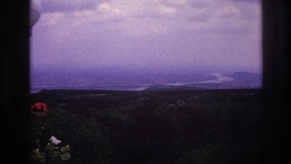 1967 France home-movie frame of a hazy valley and waterway beyond a dark window frame, with flowers in the foreground.