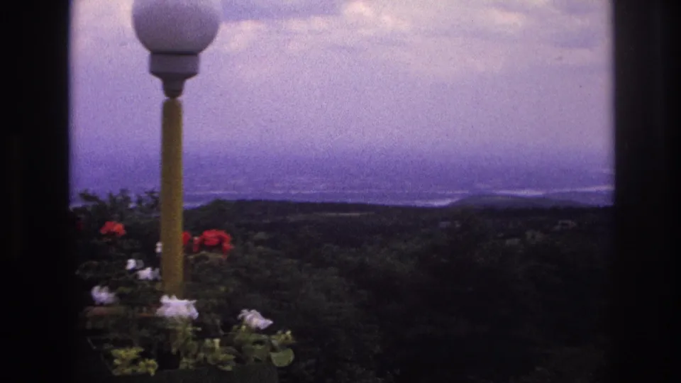 1967 France 8mm frame with a lamppost and flowering plants foregrounding a distant mountain and river-valley landscape.