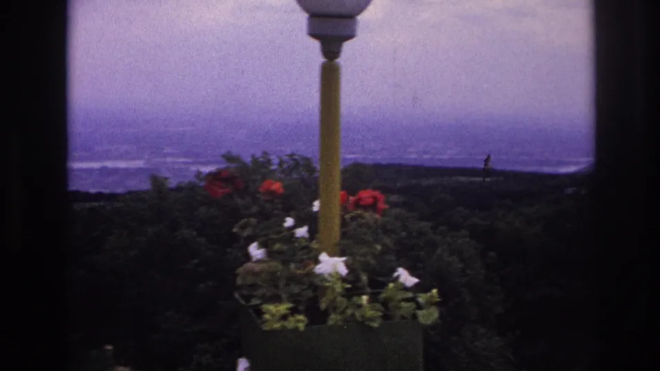 1967 France home-movie frame showing a lamppost and flowers beside a window-framed mountain and waterway view.