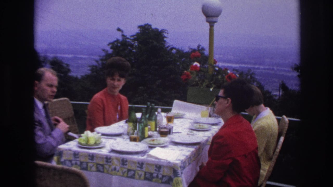 1967 France home-movie frame of four adults at a terrace dining table with bottles, plates, flowers, and a scenic hazy backdrop.