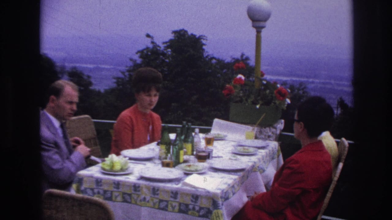 1967 France terrace dining scene with adults in 1960s clothing, table settings, bottles, flowers, and a distant landscape.