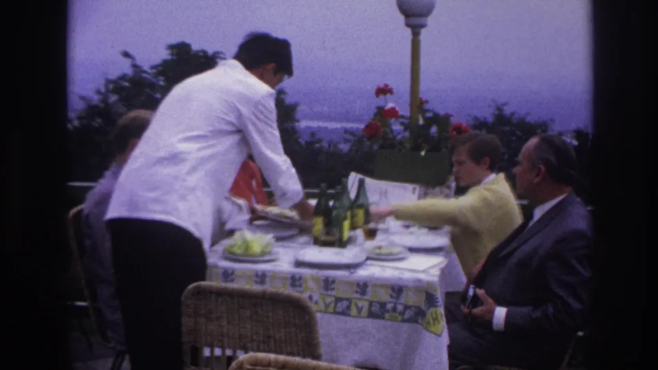 1967 France home-movie frame of a waiter serving adults at a scenic terrace table during an outdoor meal.