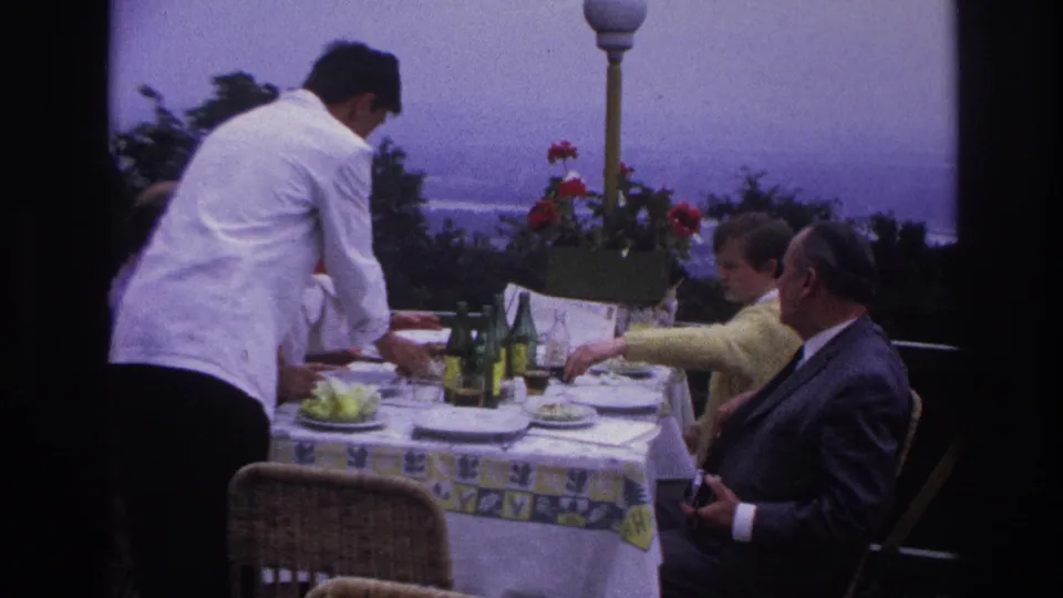 1967 France outdoor dining frame showing table service on a terrace with adults seated against a hazy landscape.