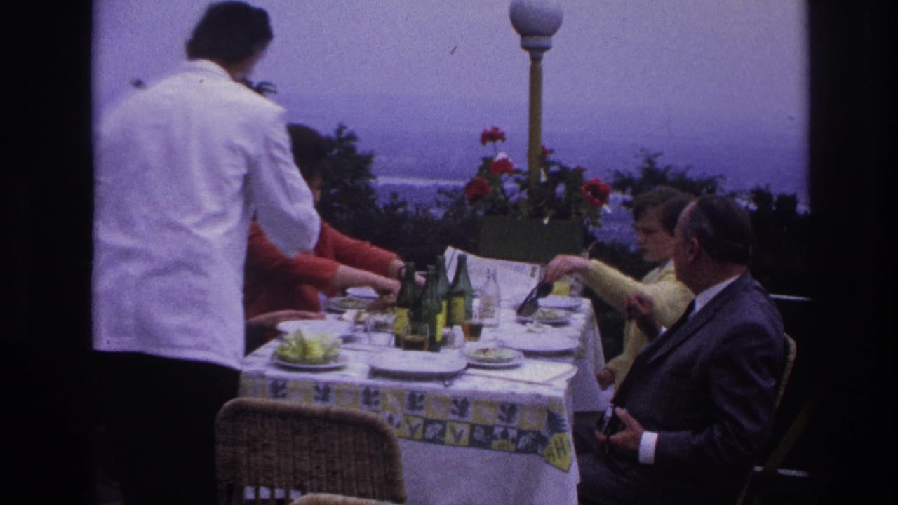In France home-movie footage from 1967, a waiter serves adults seated at a terrace lunch table with bottles and plates.