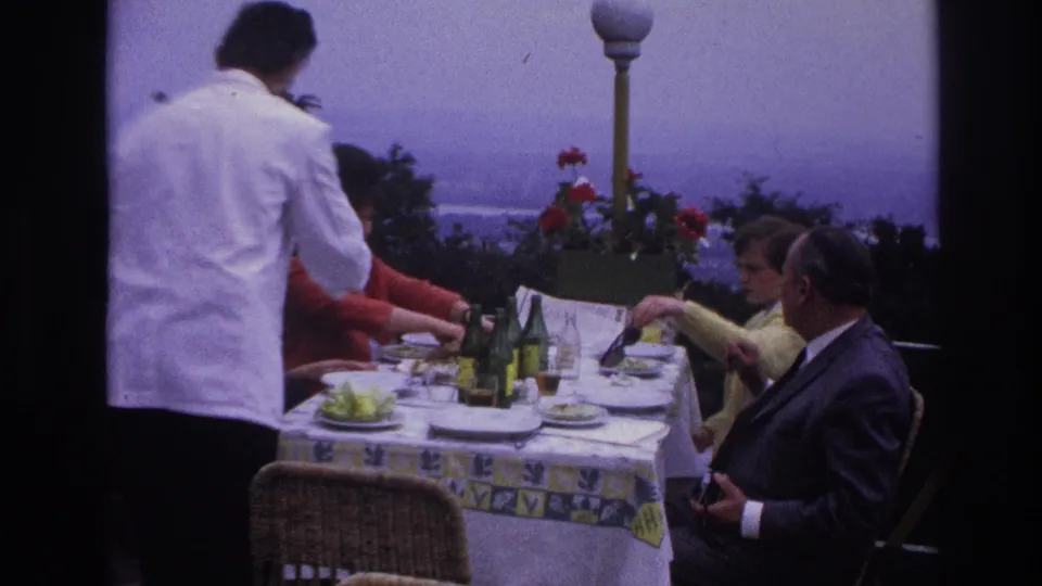 In France home-movie footage from 1967, a waiter serves adults seated at a terrace lunch table with bottles and plates.