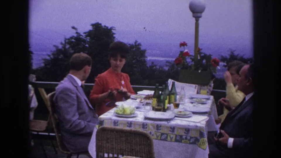A 1967 France terrace lunch scene shows adults talking around a set table with green bottles, dishes, and a scenic background.