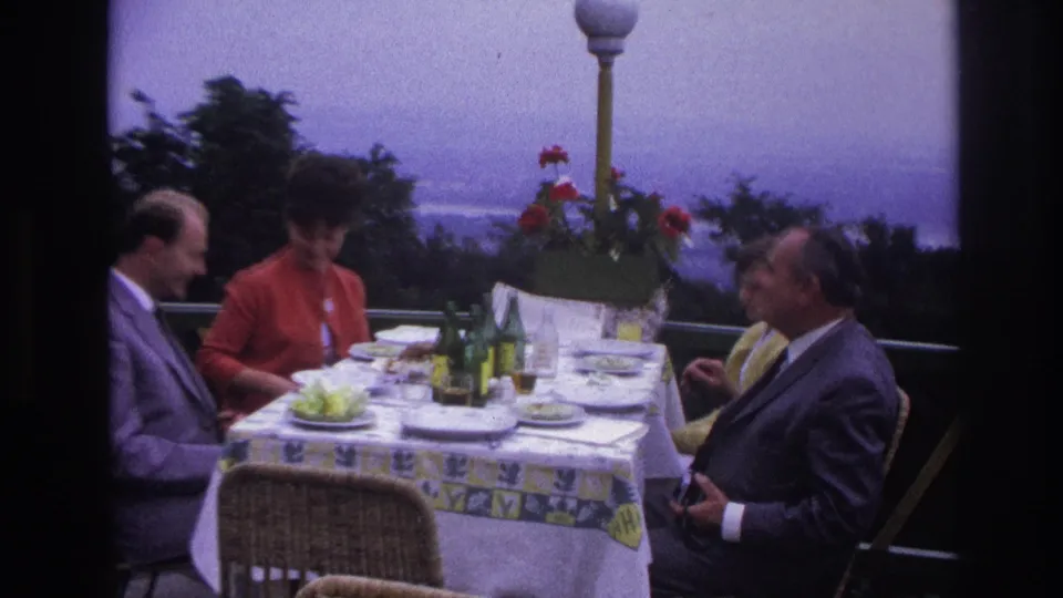 Four adults sit at a terrace lunch table in 1967 France, with bottles, plates, flowers, and a distant hazy view behind them.