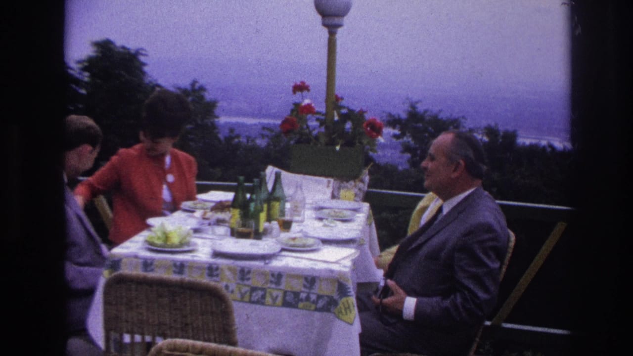 1967 France 8mm frame of adults dining on a terrace table with green bottles, plates, flowers, and a hazy coastal background.