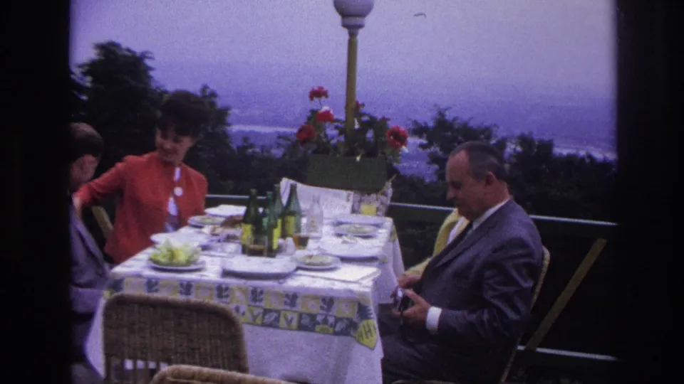 1967 France terrace dining scene with a woman in red, a man in a dark suit, table settings, bottles, and coastline beyond.