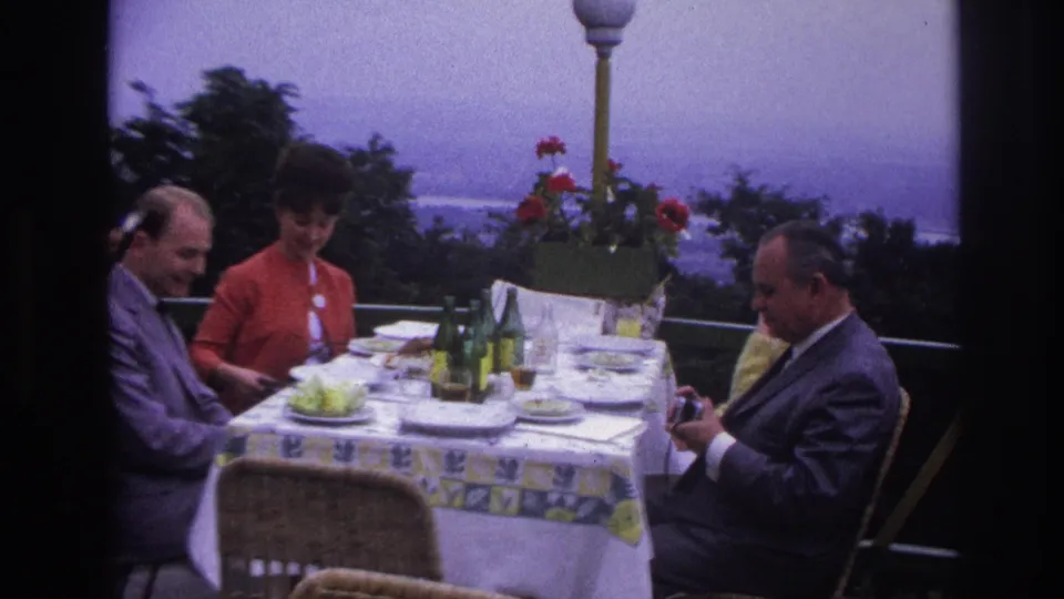 1967 France home-movie frame of three adults at an outdoor restaurant table with bottles, plates, flowers, and distant water.