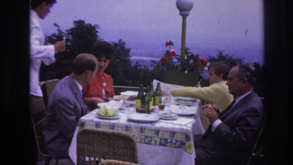 1967 France terrace restaurant frame with a waiter in white, seated diners, green bottles, plates, and coastal scenery behind.