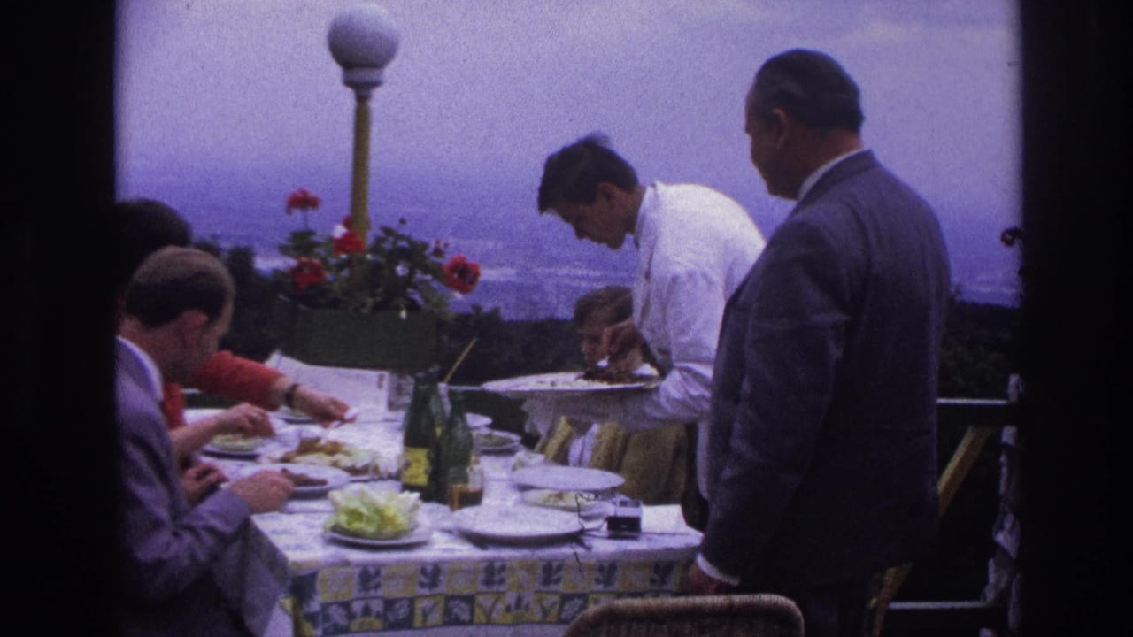 1967 France home-movie frame of adult men at a formal terrace lunch as a waiter in white serves plates beside bottles and flowers.