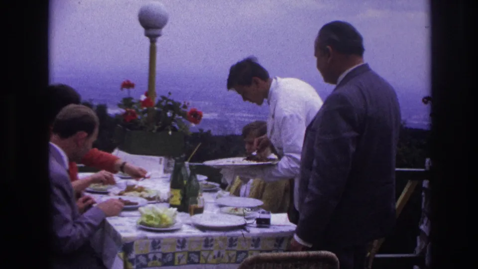 1967 France home-movie frame of adult men at a formal terrace lunch as a waiter in white serves plates beside bottles and flowers.