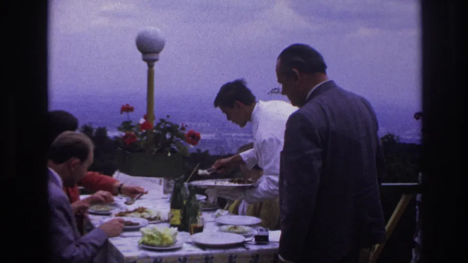 1967 France frame of a terrace meal with suited men, a waiter leaning over plates, glass bottles, red flowers, and a soft distant view.
