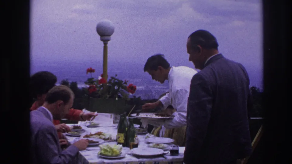 The terrace lunch continues in 1967 France, with a waiter in white serving suited guests at a patterned table overlooking a hazy distance.
