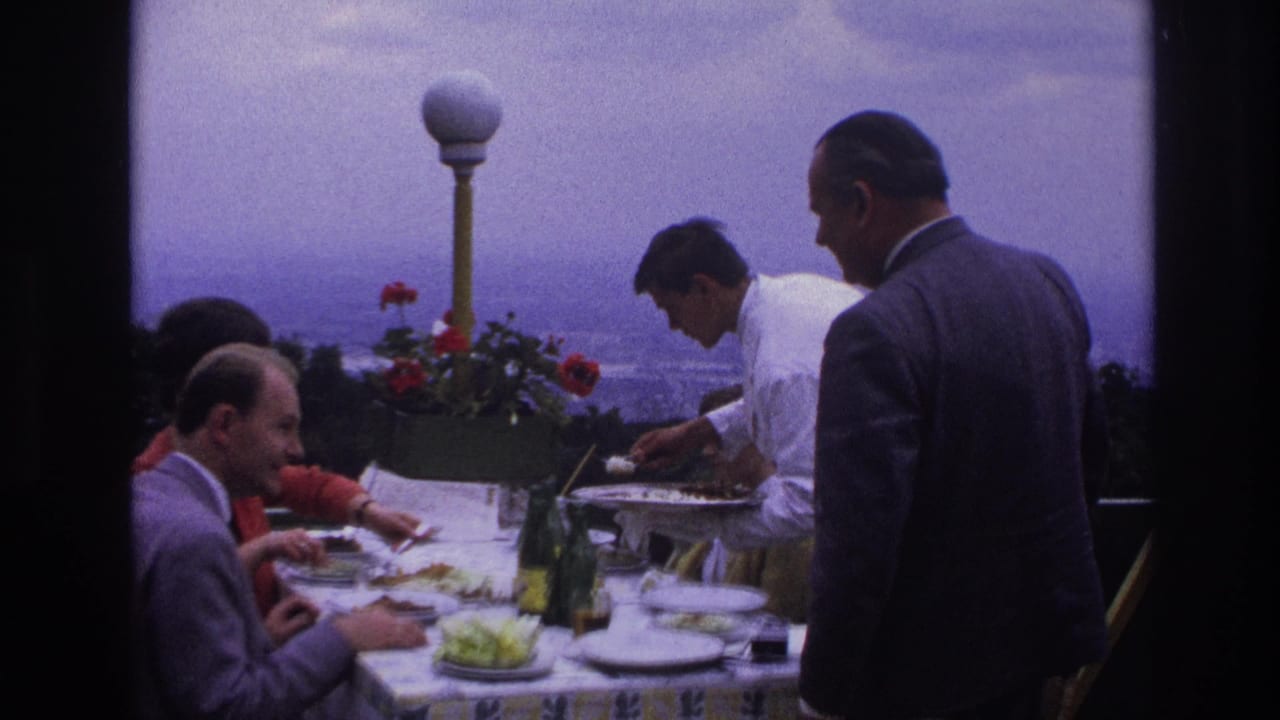 1967 home-movie frame of adults dining at a rooftop restaurant as a waiter in a white jacket serves food near flowers and a city view.